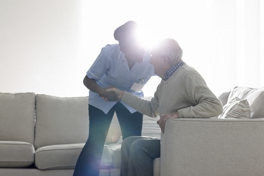 Female caregiver helping senior man get up from sofa at home