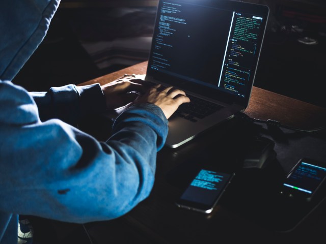 anonymous person in the hood sitting in front of computer working with laptop and mobile phone