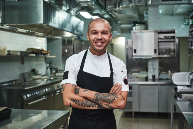 I love my work Cheerful young chef in apron keeping tattooed arms crossed and smiling while standing in a restaurant kitchen