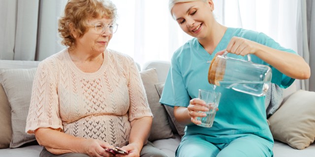 Smiling nurse pouring glass of water for elderly woman on sofa