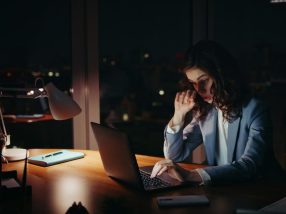Sleepy businesswoman working night sitting at office desk. Lady looking laptop.