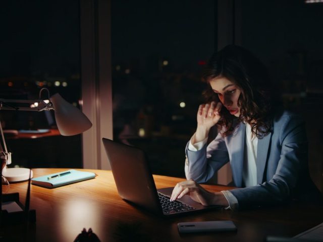 Sleepy businesswoman working night sitting at office desk. Lady looking laptop.